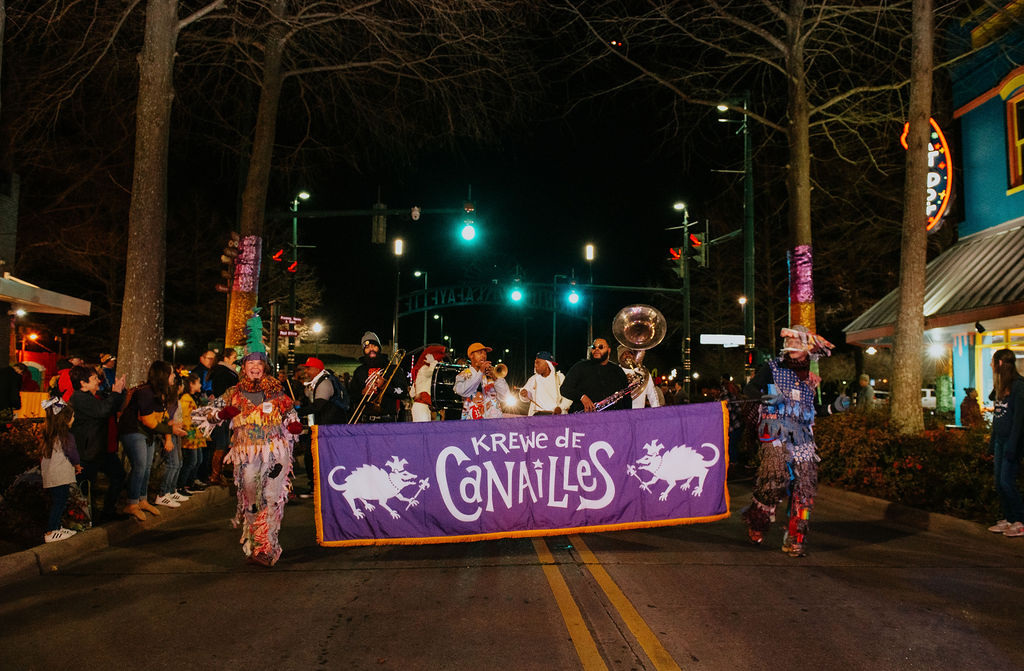 Photo of costumed Krewe de Canailles members marching in downtown Lafayette, Louisiana while holding the Krewe de Canailles banner and playing instruments.